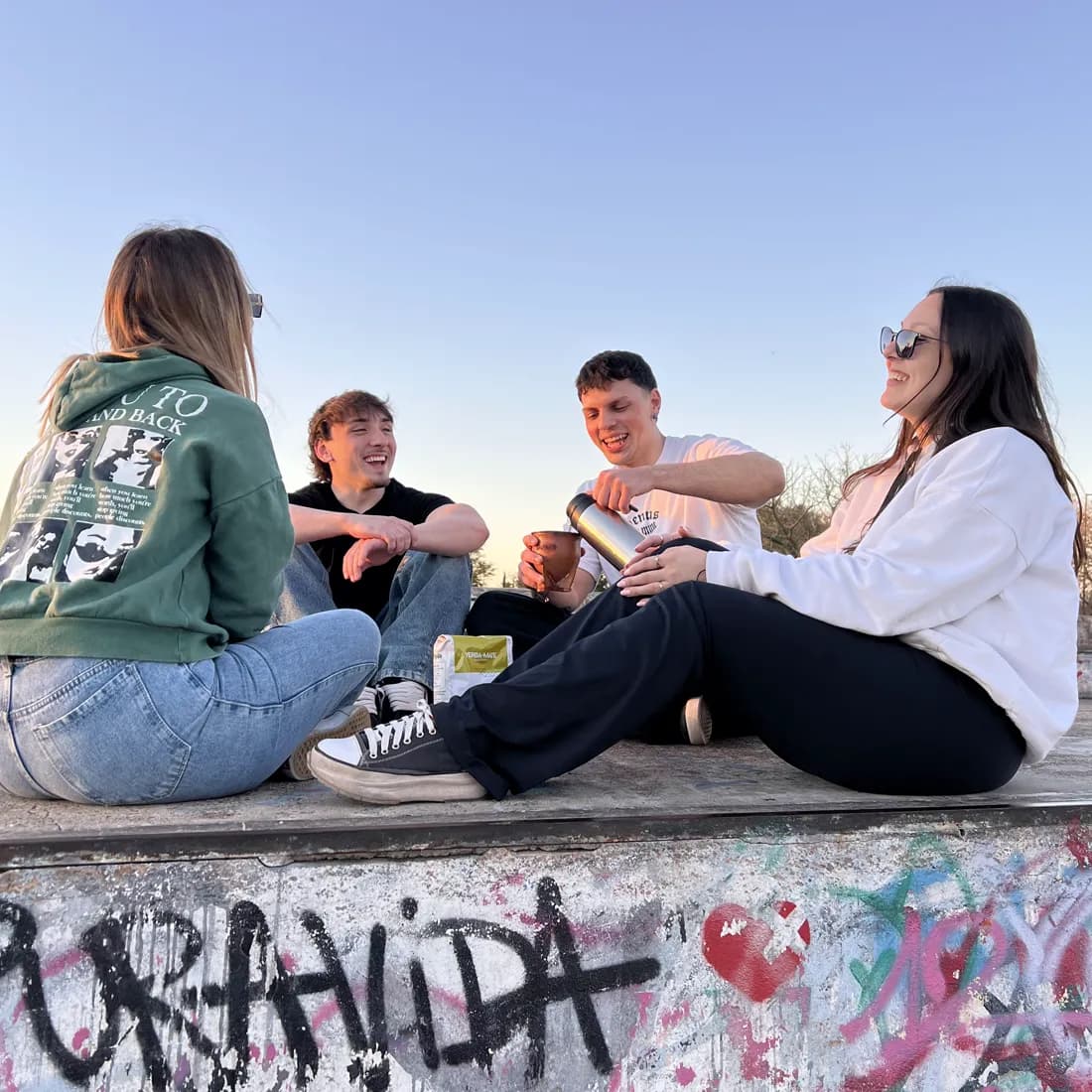 Cuatro amigos sentados en una rampa de skate al atardecer, cebando un mate, con un paquete de LaTiNa apoyado al lado